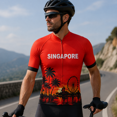 A man wearing a red cycling jersey with a tropical sunset and palm tree design, black cycling shorts, a black helmet, and sunglasses, riding a road bike on a scenic mountain road.
