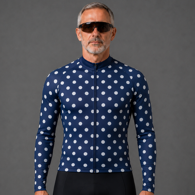 A man wearing a navy blue long-sleeve cycling jersey with a white polka-dot pattern and black cycling shorts, standing against a solid gray studio background.