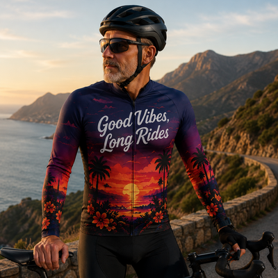A man wearing a long-sleeve cycling jersey with a sunset tropical print and the text 'Good Vibes, Long Rides' stands on a coastal road with a mountain backdrop, holding a road bike.