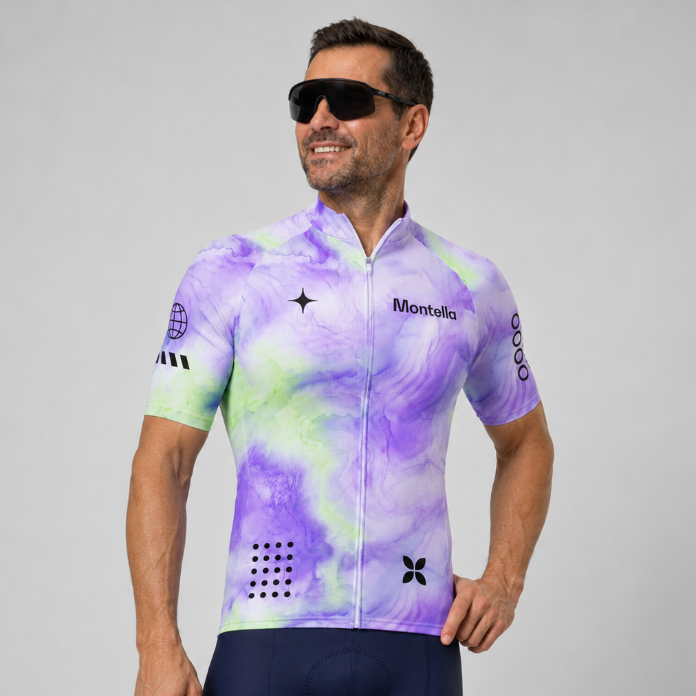 A man wearing a short-sleeve cycling jersey with a purple, green, and white tie-dye pattern, paired with navy blue cycling shorts and black sunglasses, against a solid gray studio background.